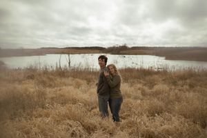 Two people share a tender moment while standing in tall grass near a lake under cloudy skies.