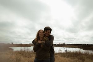 A loving couple embraces while standing near a lake on an overcast autumn day.