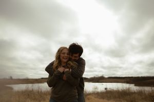 Two people share a romantic embrace in front of a lake on a cloudy day.