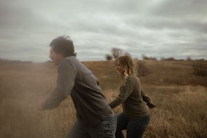 A playful couple runs through tall grass on an overcast day near a lake.