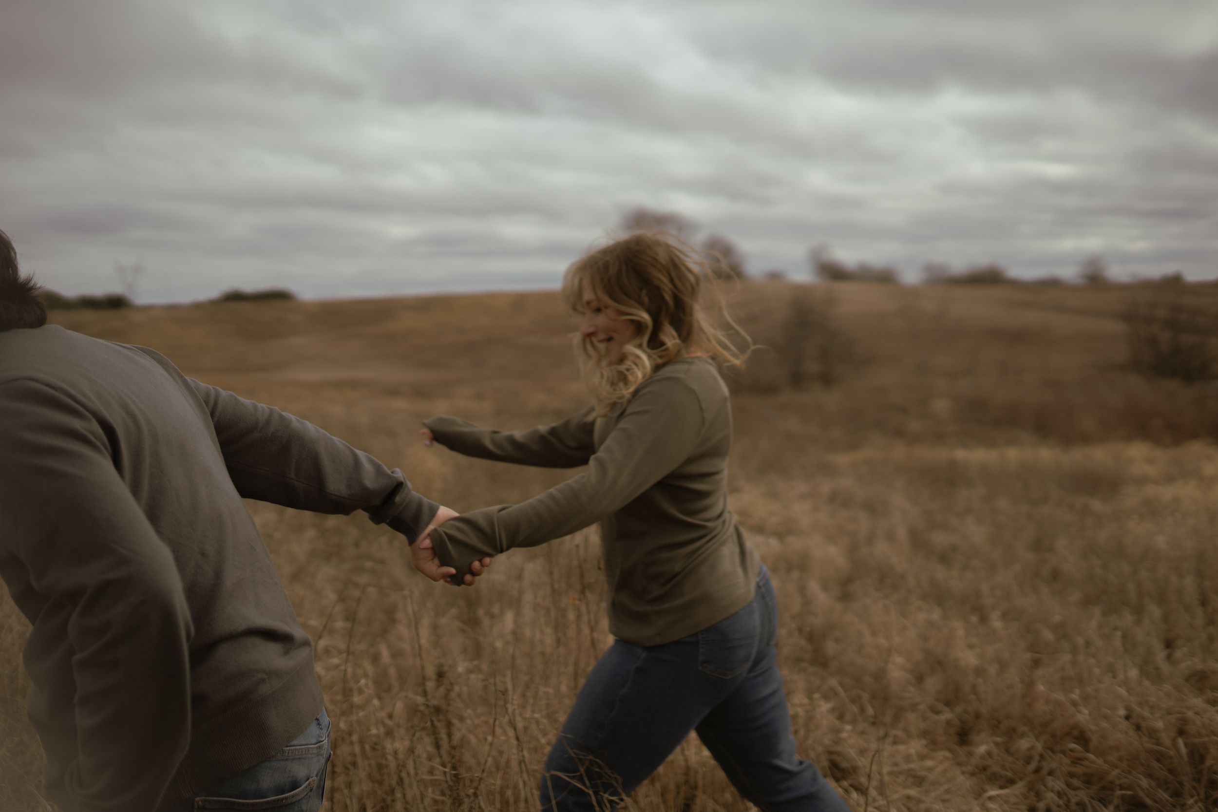 Two people playfully interact in a golden field under cloudy skies.