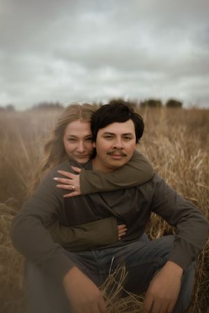 Playful couple sitting in tall grass field during overcast day.