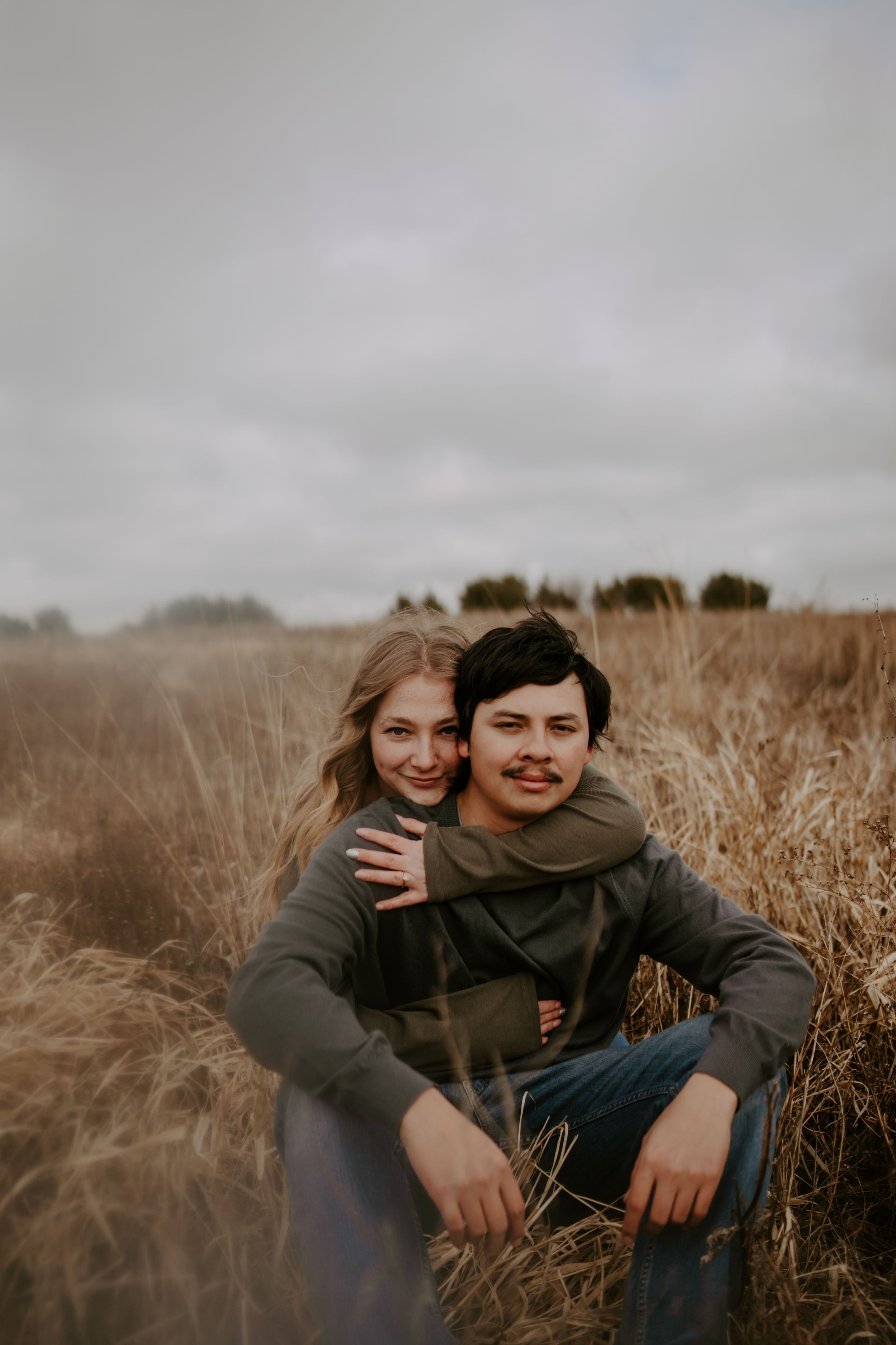 Couple laughing together while sitting in tall autumn grass under cloudy skies.