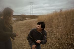 Two people interact playfully on a dirt path surrounded by dried grass and utility poles.