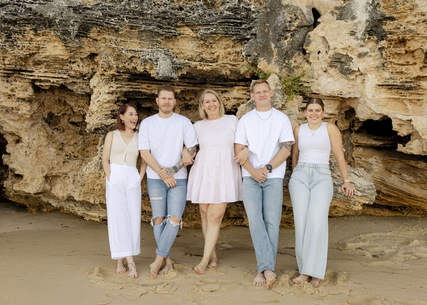A family group poses for beach portraits against a rocky coastal backdrop in coordinated casual outfits.