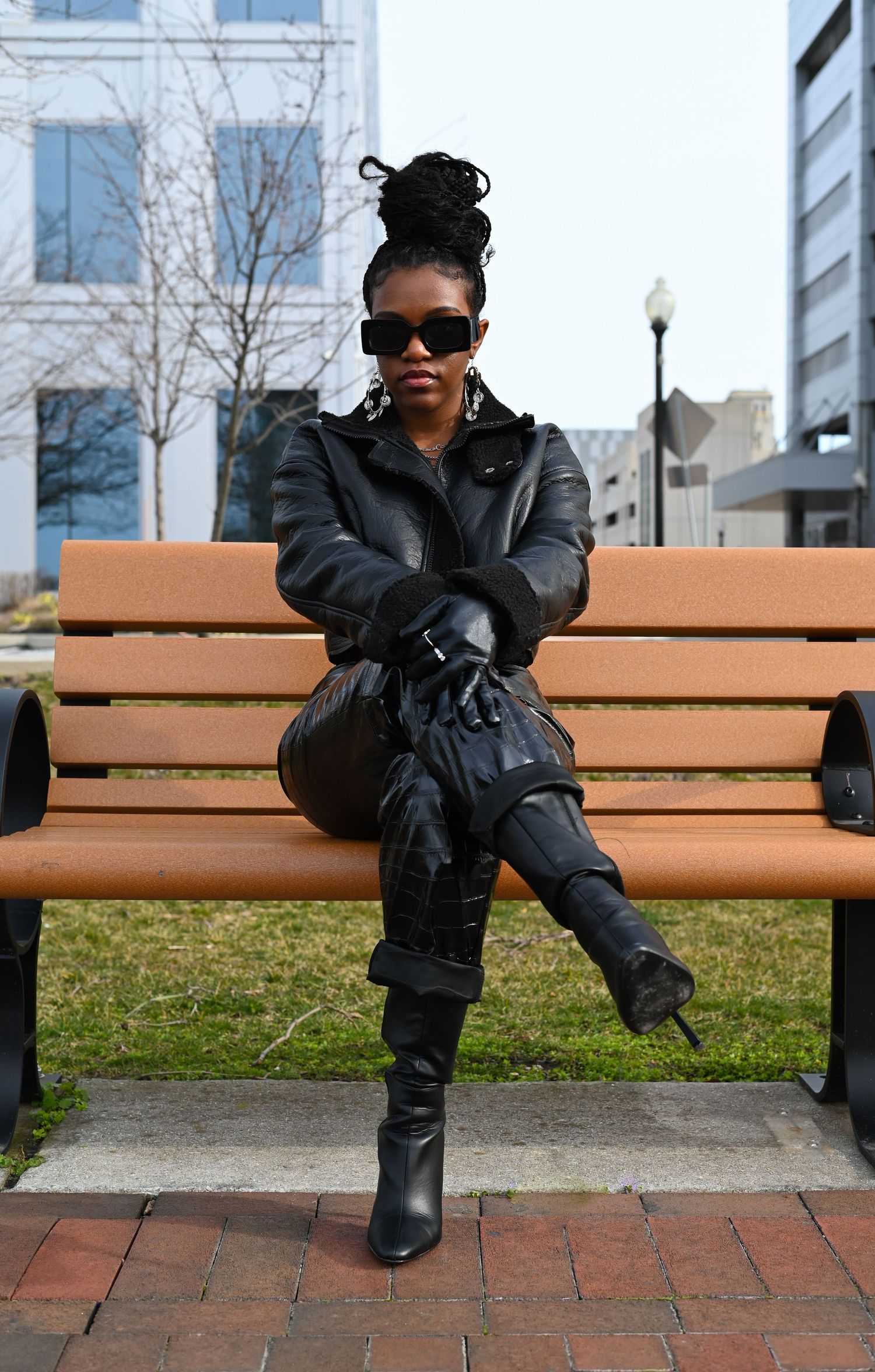 Stylish person in black leather jacket and boots sitting confidently on wooden bench in urban setting.
