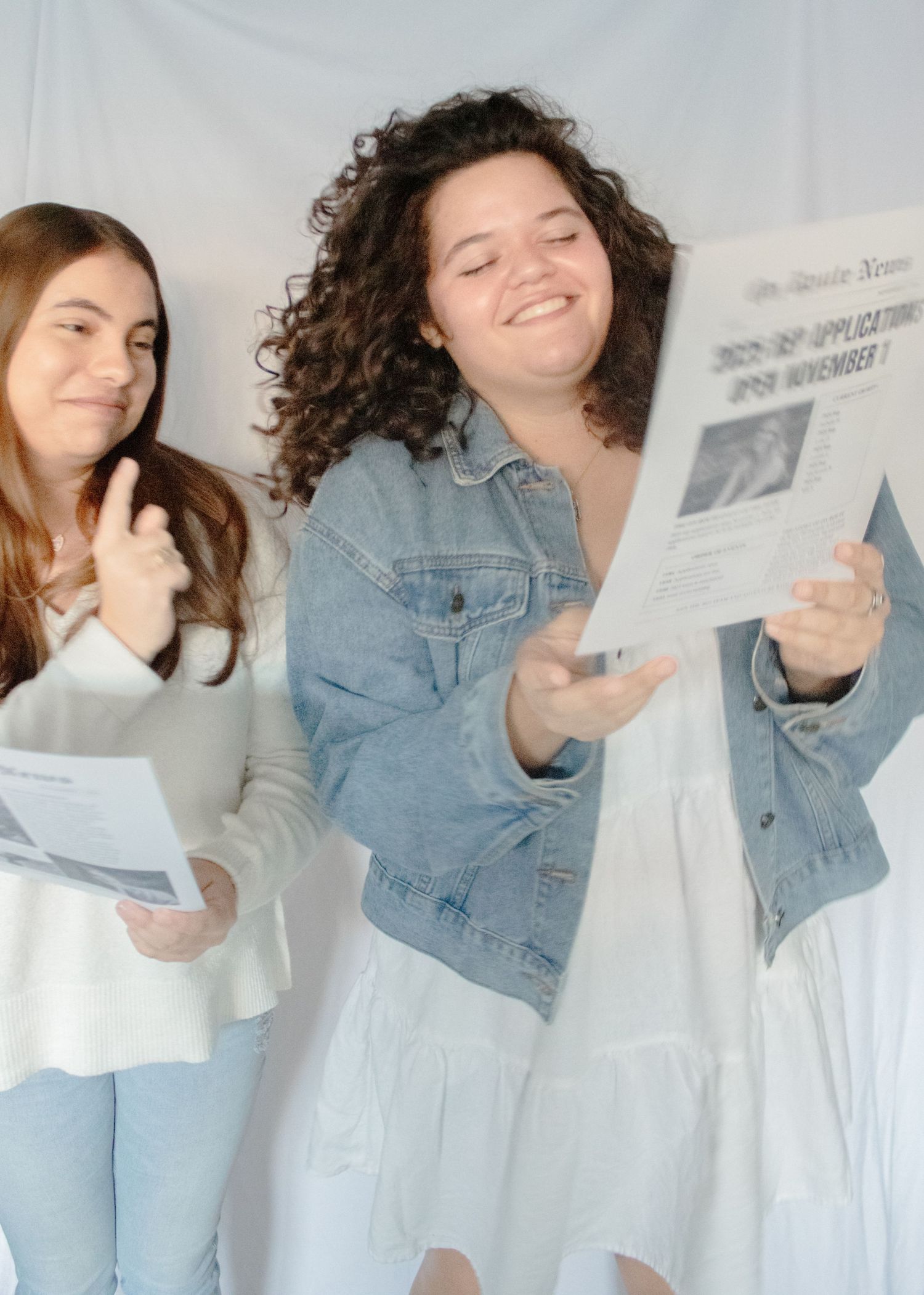 Two people in casual attire reading and laughing together while holding papers against a white background.