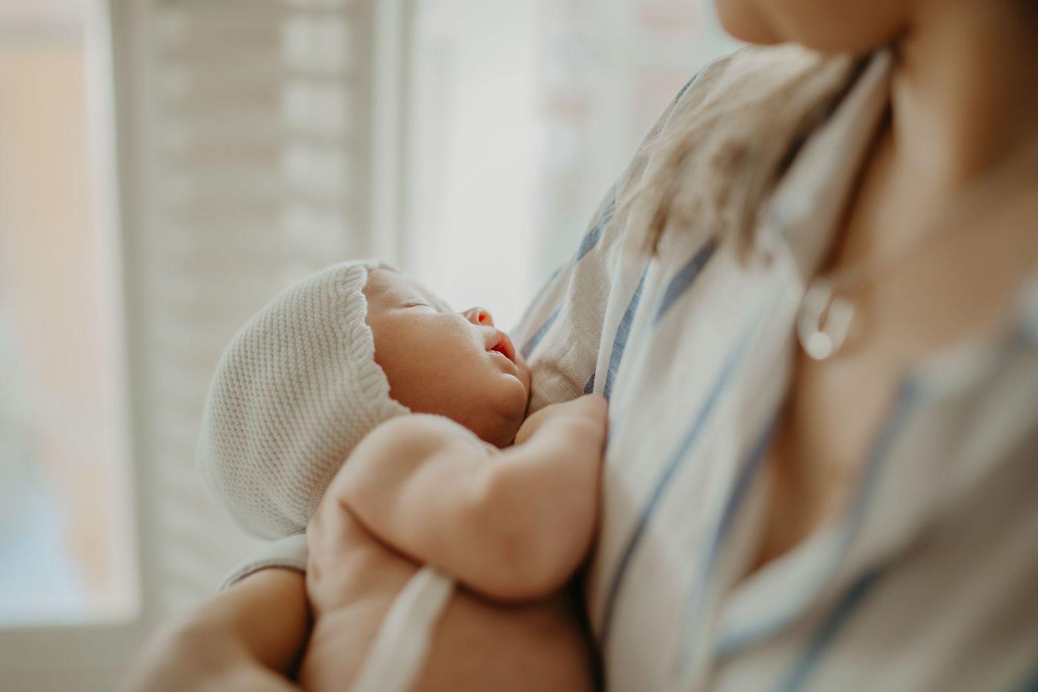 Mother gently holding and nursing newborn baby in warm natural light near window.