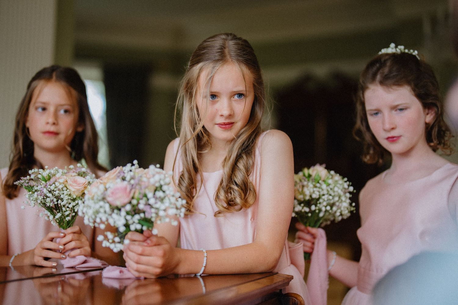 Young flower girl in pink dresses hold small white and pink bouquets while leaning against a piano