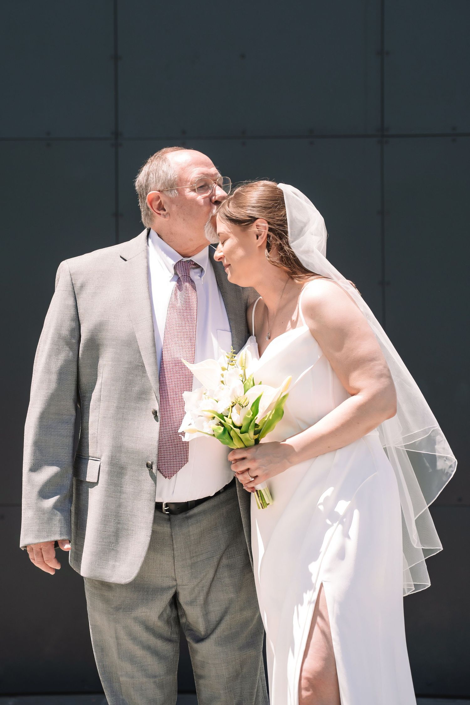 A tender wedding moment between two people in formal attire, with the bride holding a white lily bouquet against a gray wall.