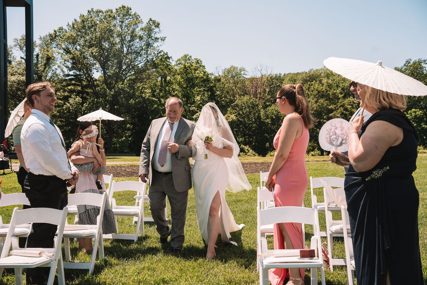 A small outdoor wedding ceremony takes place on a sunny day with white chairs set up on grass with trees in background.