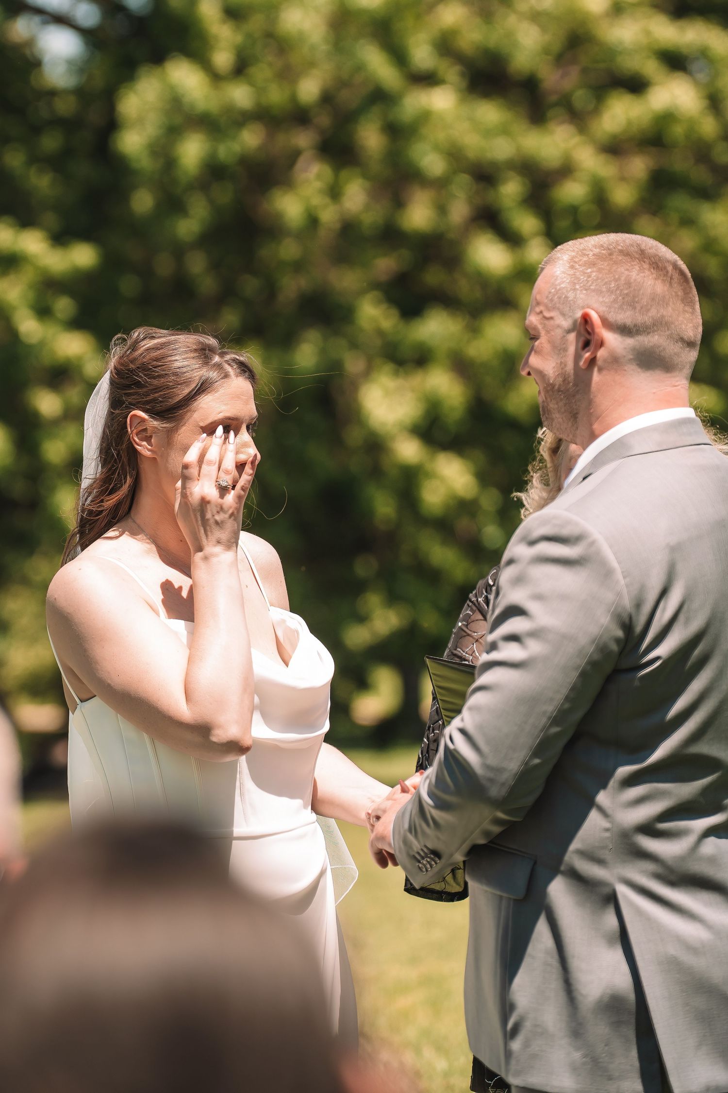 A touching outdoor wedding ceremony moment as bride covers her face with emotion while holding hands with groom.