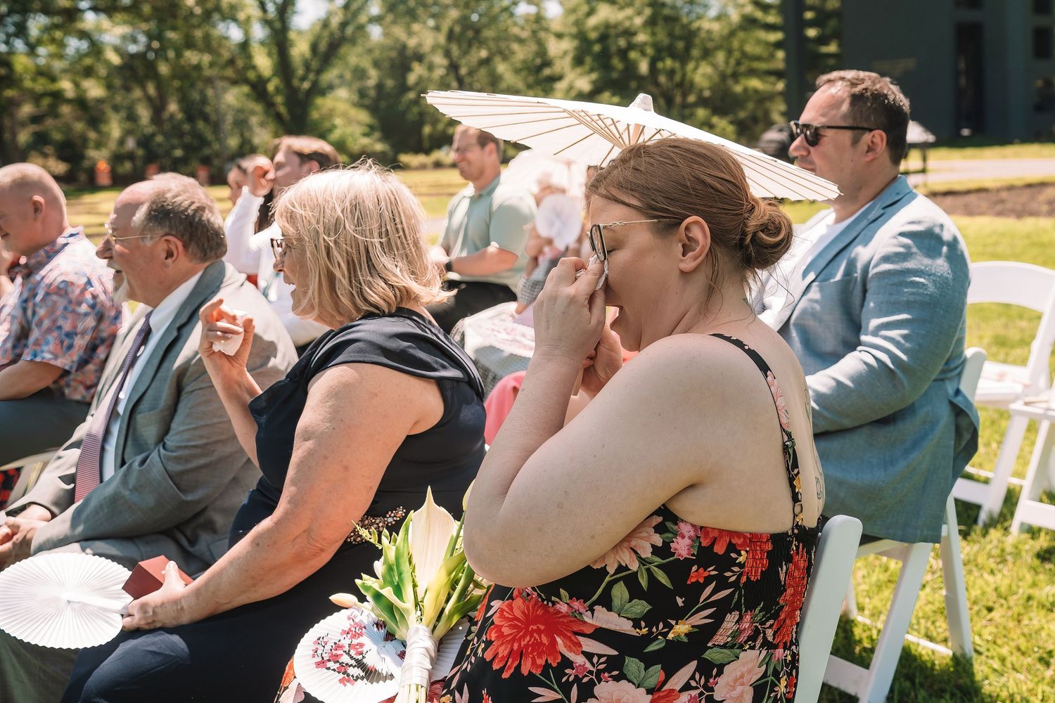 Guests seated in white chairs at an outdoor summer wedding ceremony react emotionally in the sunlight.