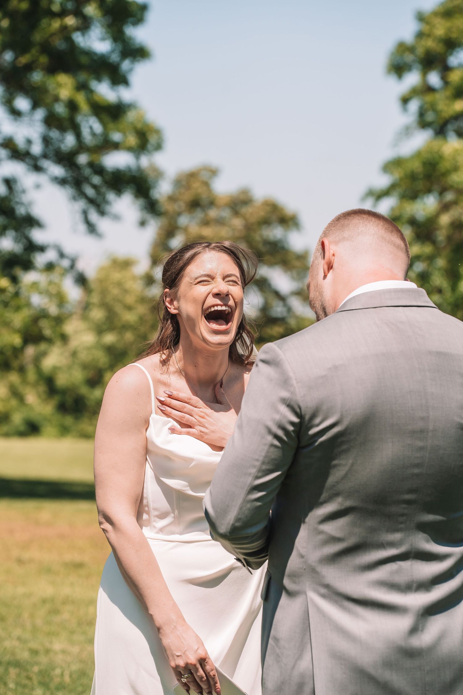 A person in a white wedding dress laughs joyfully while sharing a moment with someone in a grey suit outdoors.