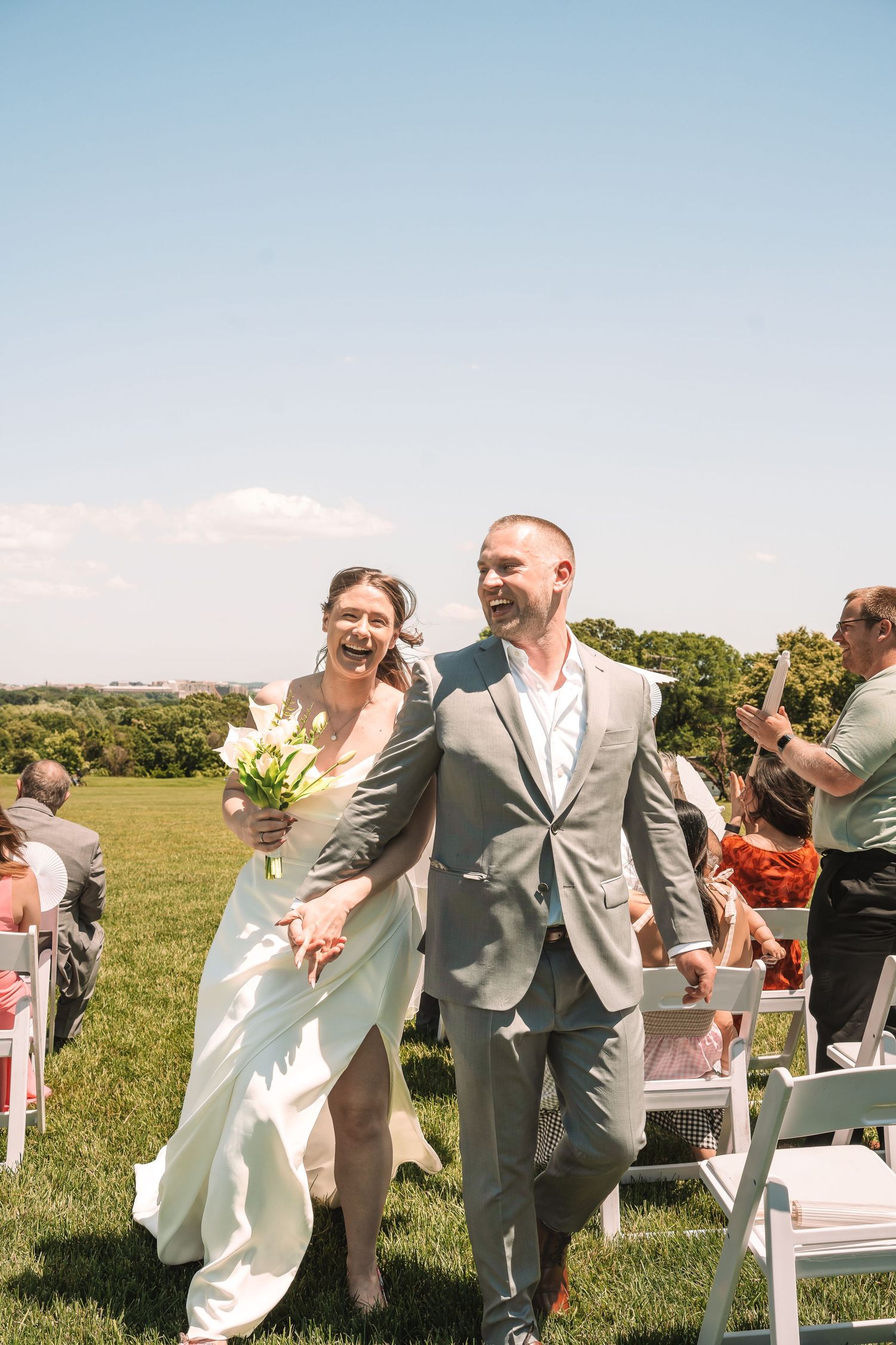 A joyful couple walks down the outdoor aisle together after their wedding ceremony on a sunny day in a grassy field.