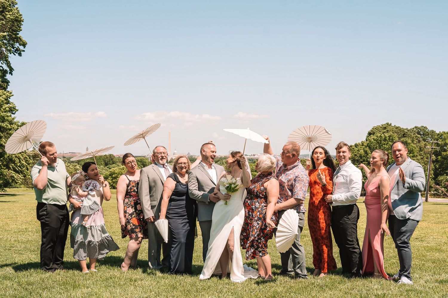 Wedding party guests pose together outdoors on a sunny day with green grass and trees in the background.