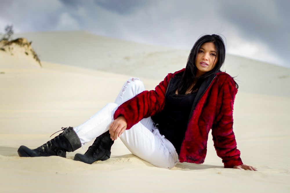 A person in a red velvet jacket and white pants reclines on sand dunes under a cloudy sky.