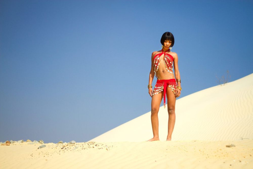 Person wearing red shorts and tank top stands on top of bright sand dune against clear blue sky.