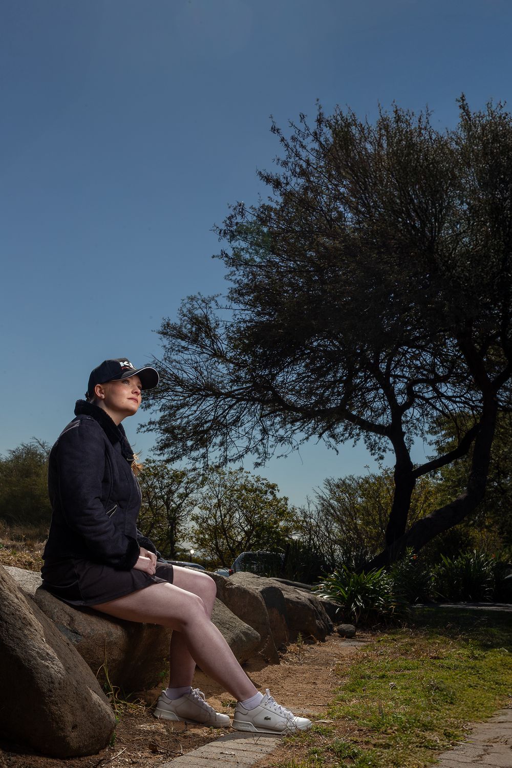 Someone in casual attire sits on large rocks along a hiking trail beneath a gnarled tree against a bright blue sky.