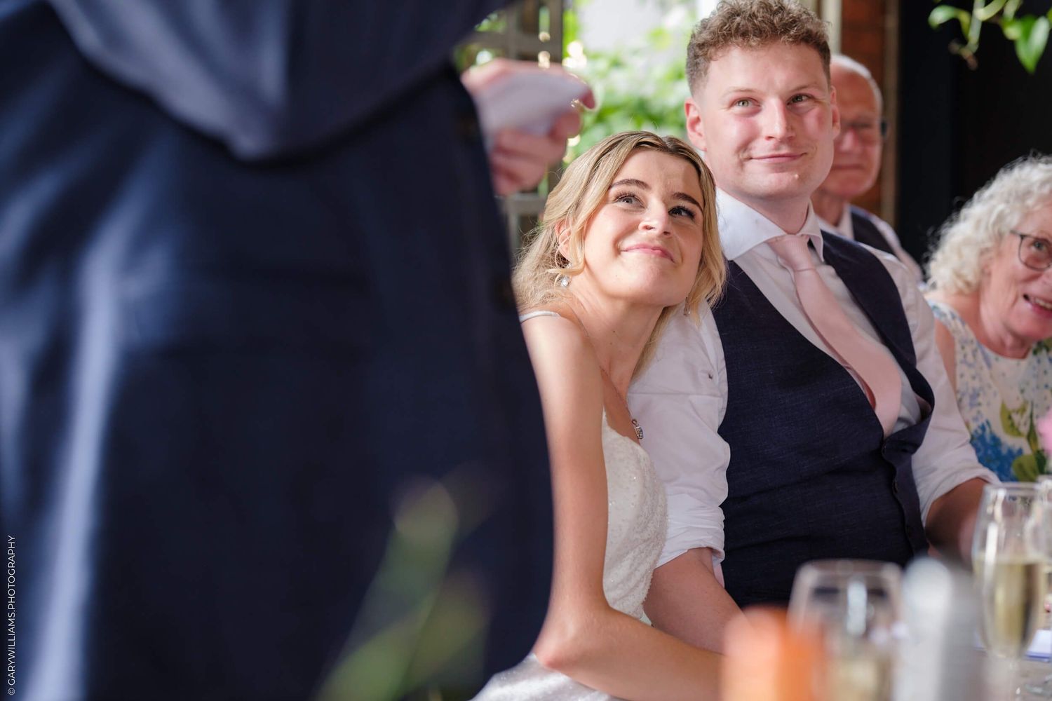 Wedding guests seated at reception tables smile and laugh during speeches at an indoor celebration.