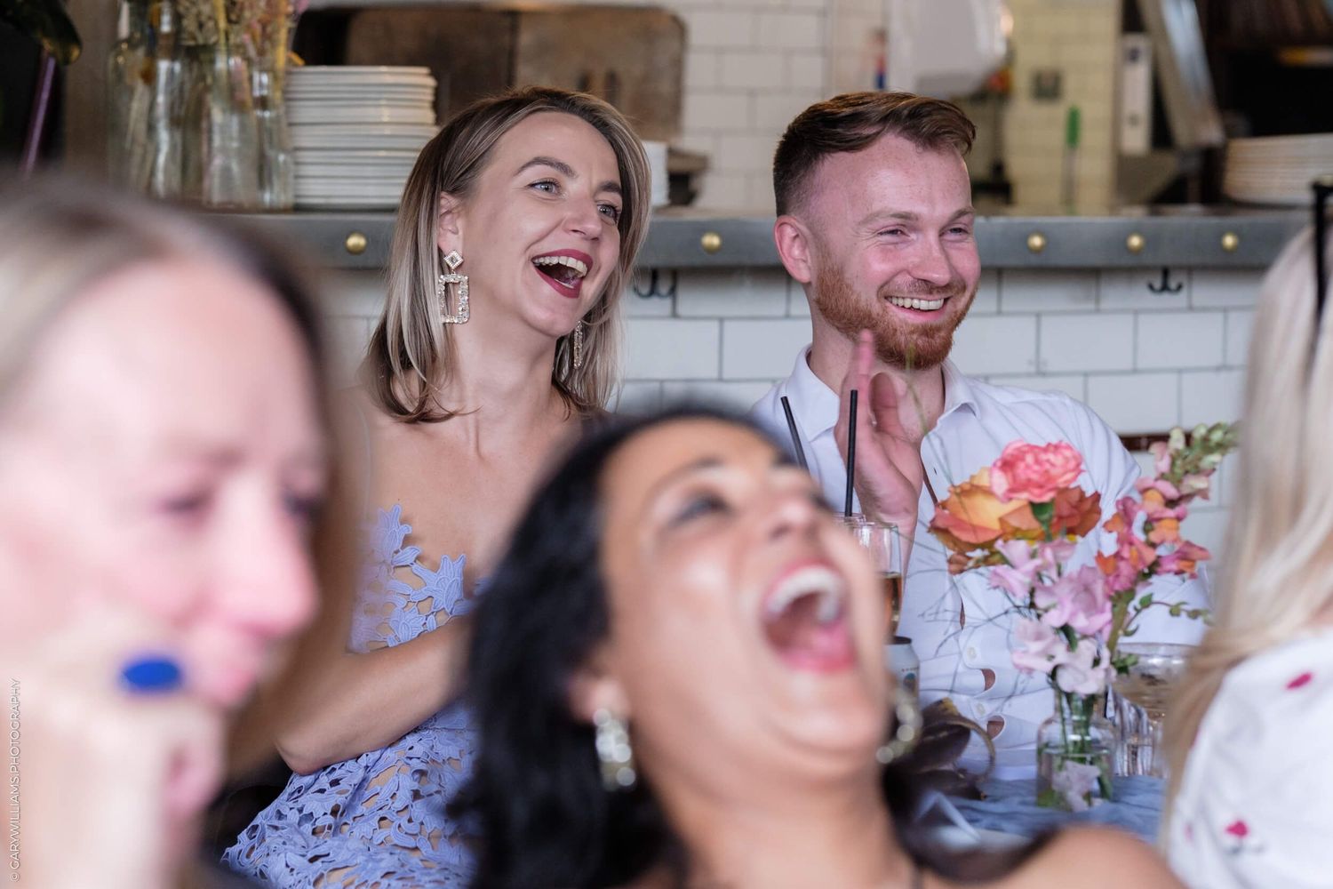 Happy guests sharing laughter and joy during a celebratory gathering with drinks and flowers on the table.