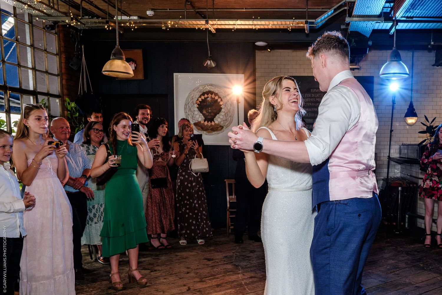 Wedding guests dance together on a rustic wooden dance floor under warm lighting at an indoor reception venue.