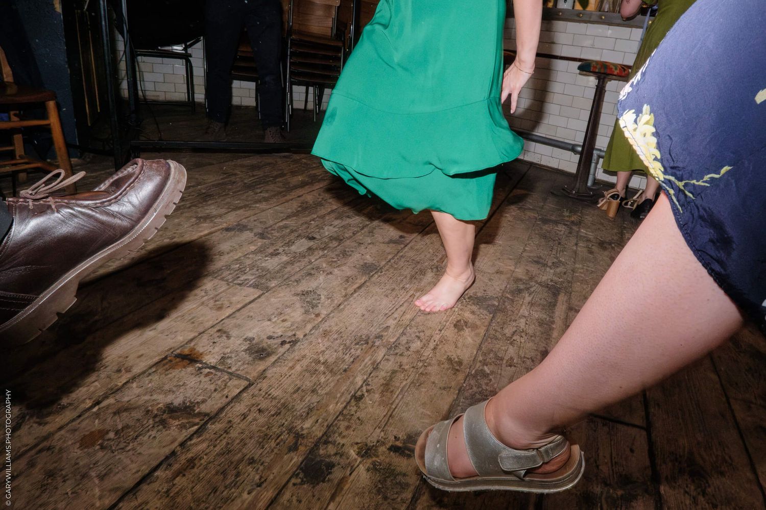 A person in a green dress stands barefoot on old wooden floorboards next to others wearing shoes.