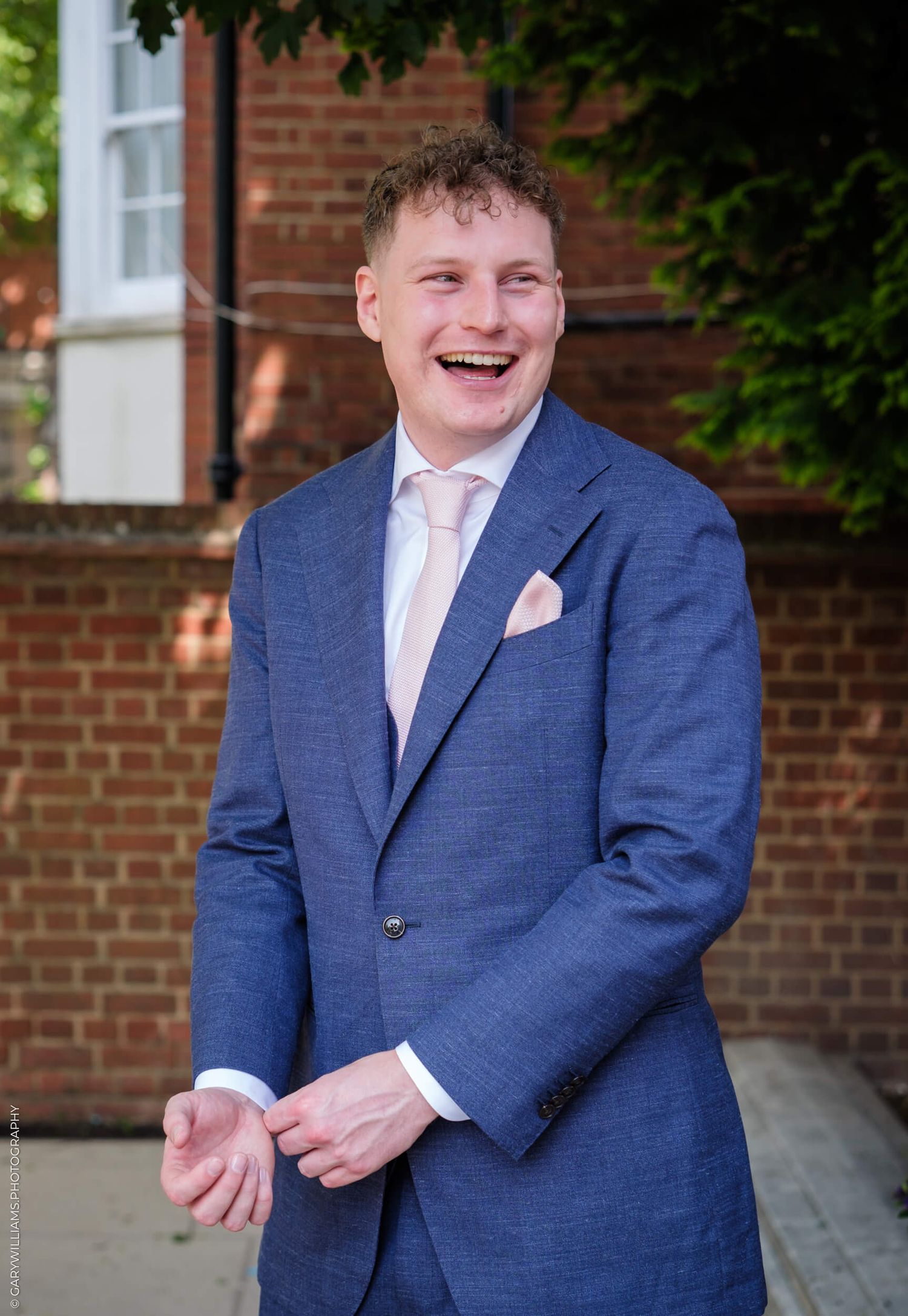 A person wearing a navy blue suit and pale pink tie smiles warmly in front of a brick building.