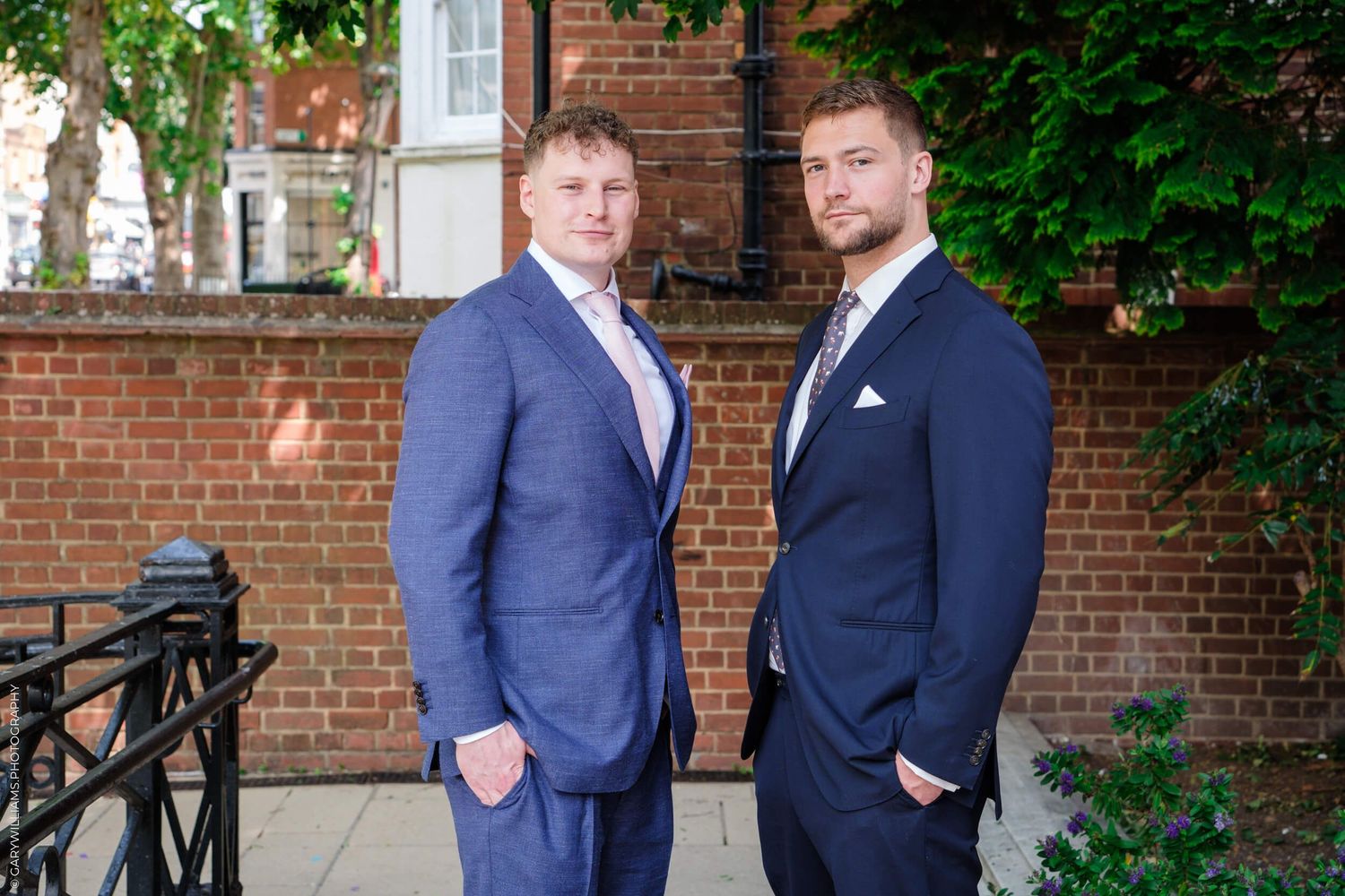 Two people in navy blue suits standing together in front of a brick building with green foliage.