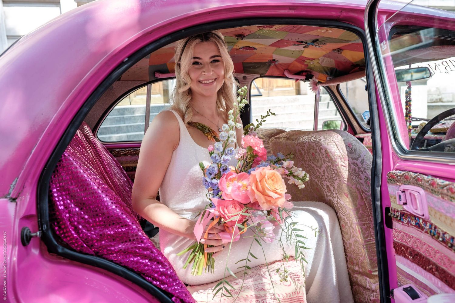 Person in white dress holding colorful bouquet sits inside bright pink vintage car with custom interior.