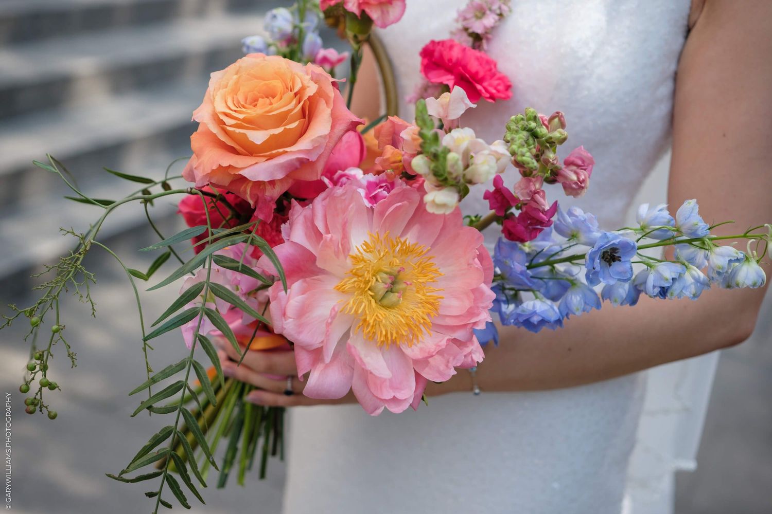 Close-up of a vibrant bridal bouquet featuring coral roses, pink peonies, and delicate blue flowers with greenery.