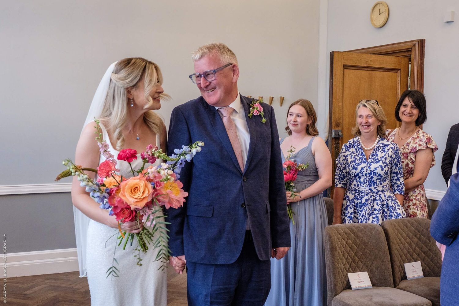 A wedding party smiles in a rustic indoor venue with colorful floral bouquets and dresses in blue and gray.