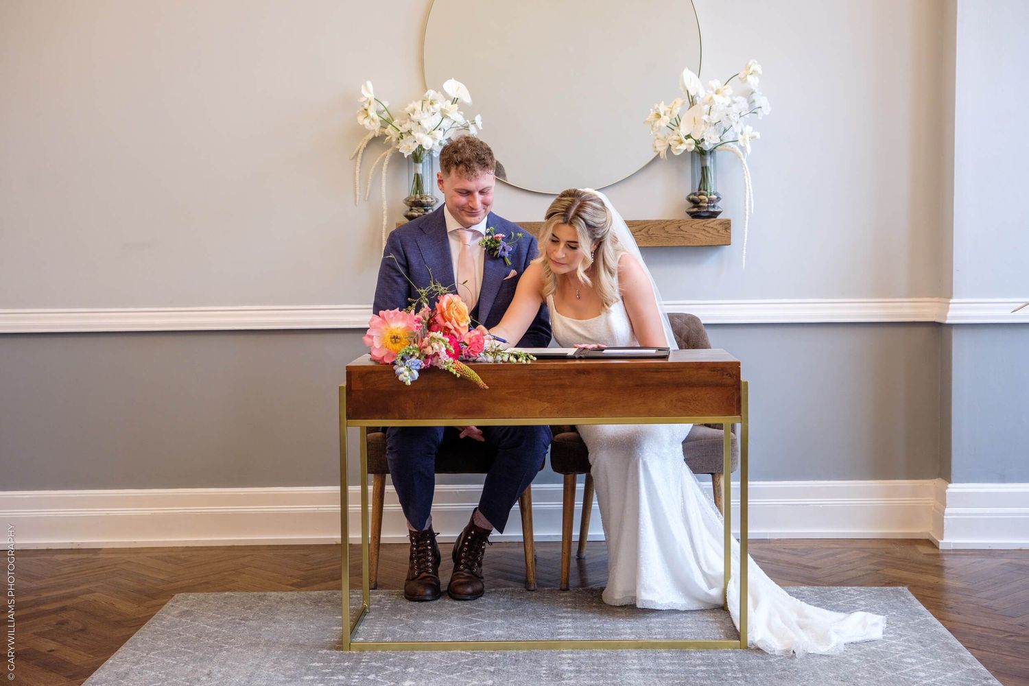 Elegant wedding scene with couple sitting at decorative table under floral hoop arch in bright indoor venue.