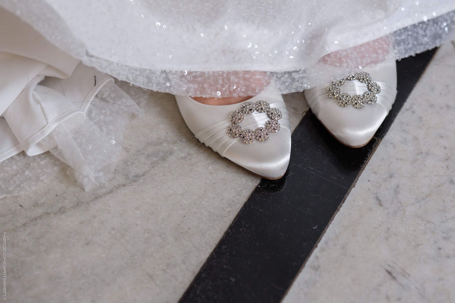 Elegant white bridal shoes with crystal embellishments peek out from beneath a lacy wedding dress hem on a grey floor.