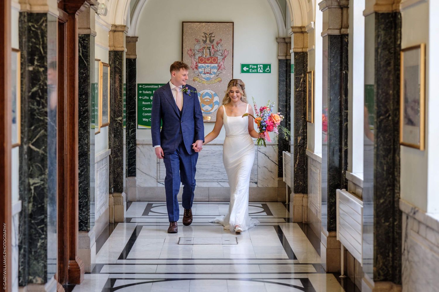 A couple walks down an elegant marble-floored hallway with ornate columns and architectural details in a historic building.