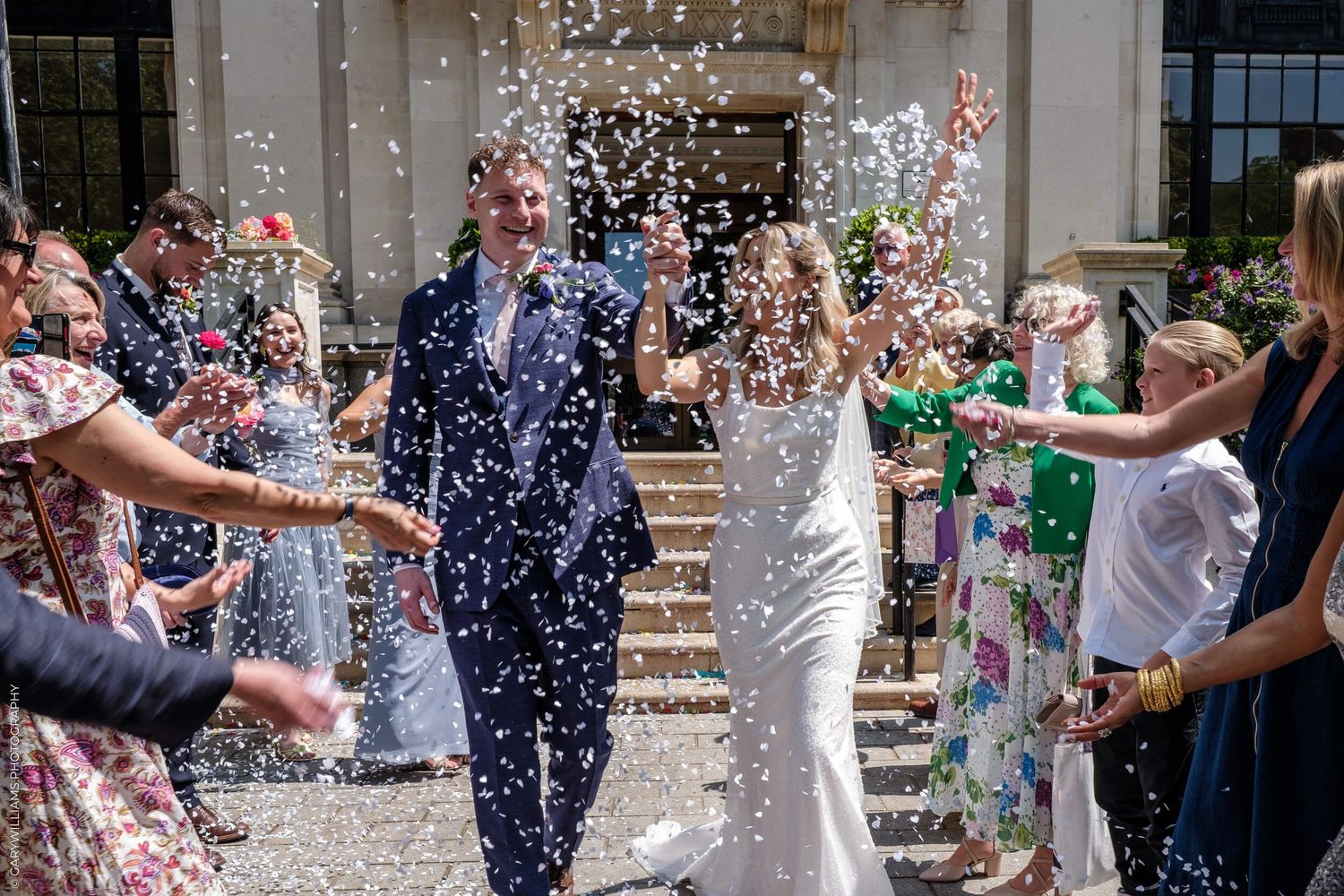 Happy wedding guests throw pink flower petals in celebration during outdoor ceremony exit.