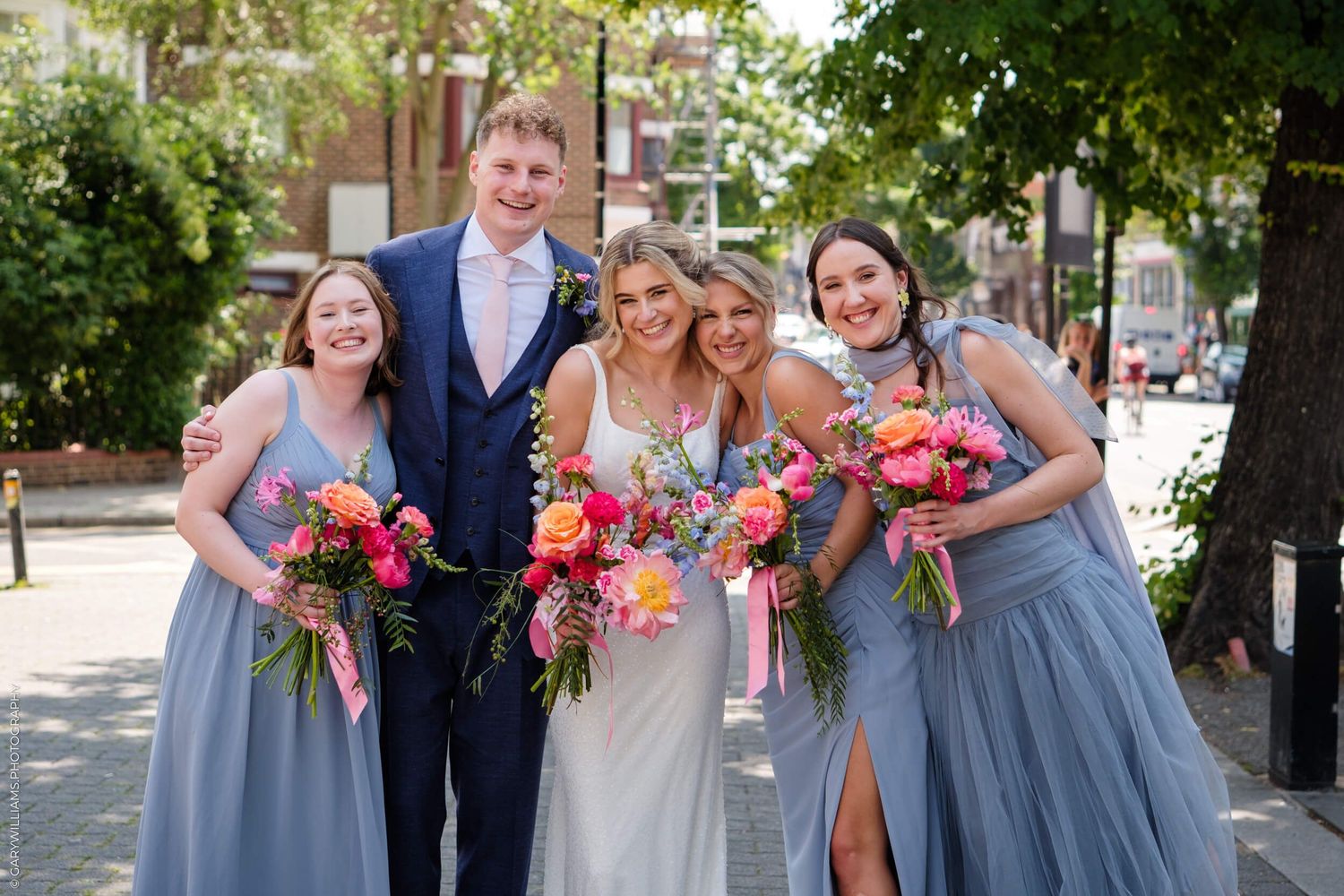 Wedding party in blue and gray formal attire poses outdoors with bright pink floral bouquets in summer.