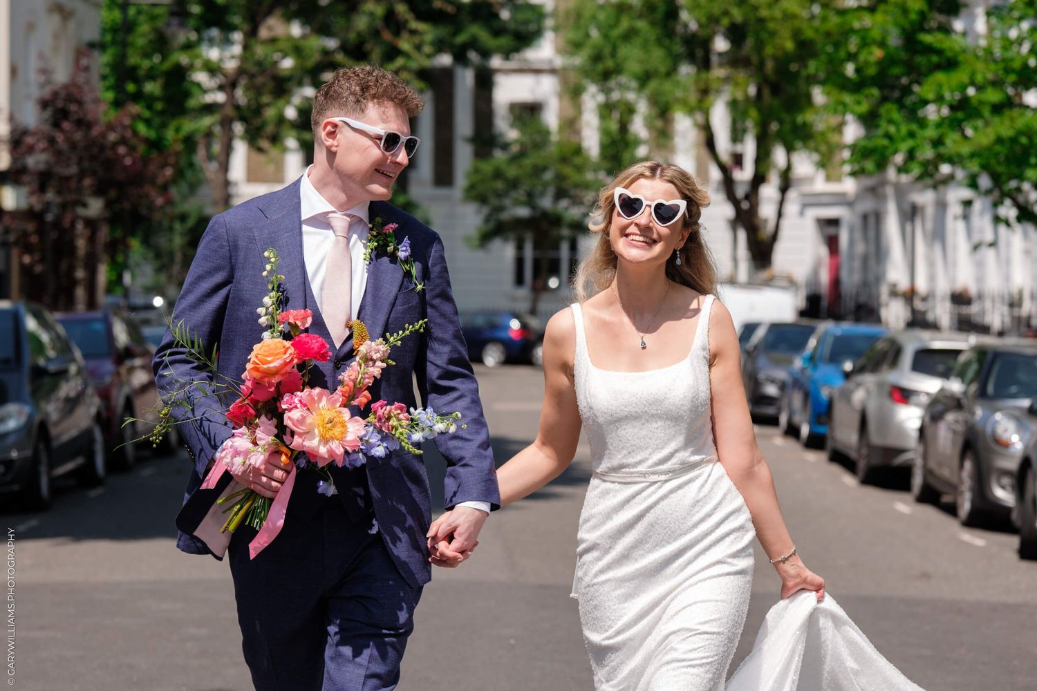 Bride in a white dress and groom in navy suit walk along urban street holding colorful floral bouquets.