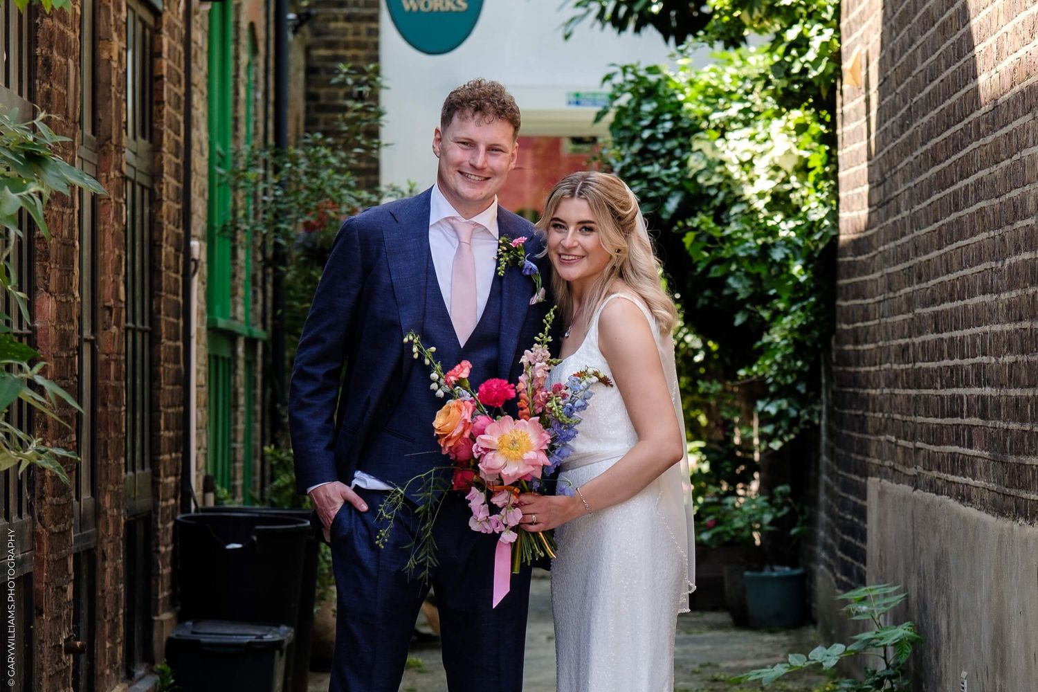 A newlywed couple stands together in an alley holding a vibrant pink and orange wedding bouquet.