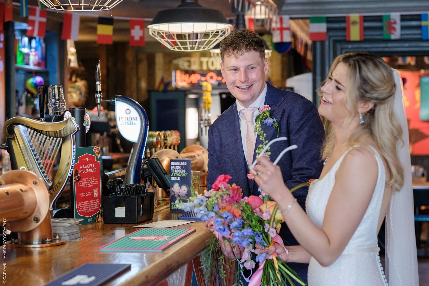 A couple celebrate at a pub bar counter with colorful flowers and beer taps visible in the background.