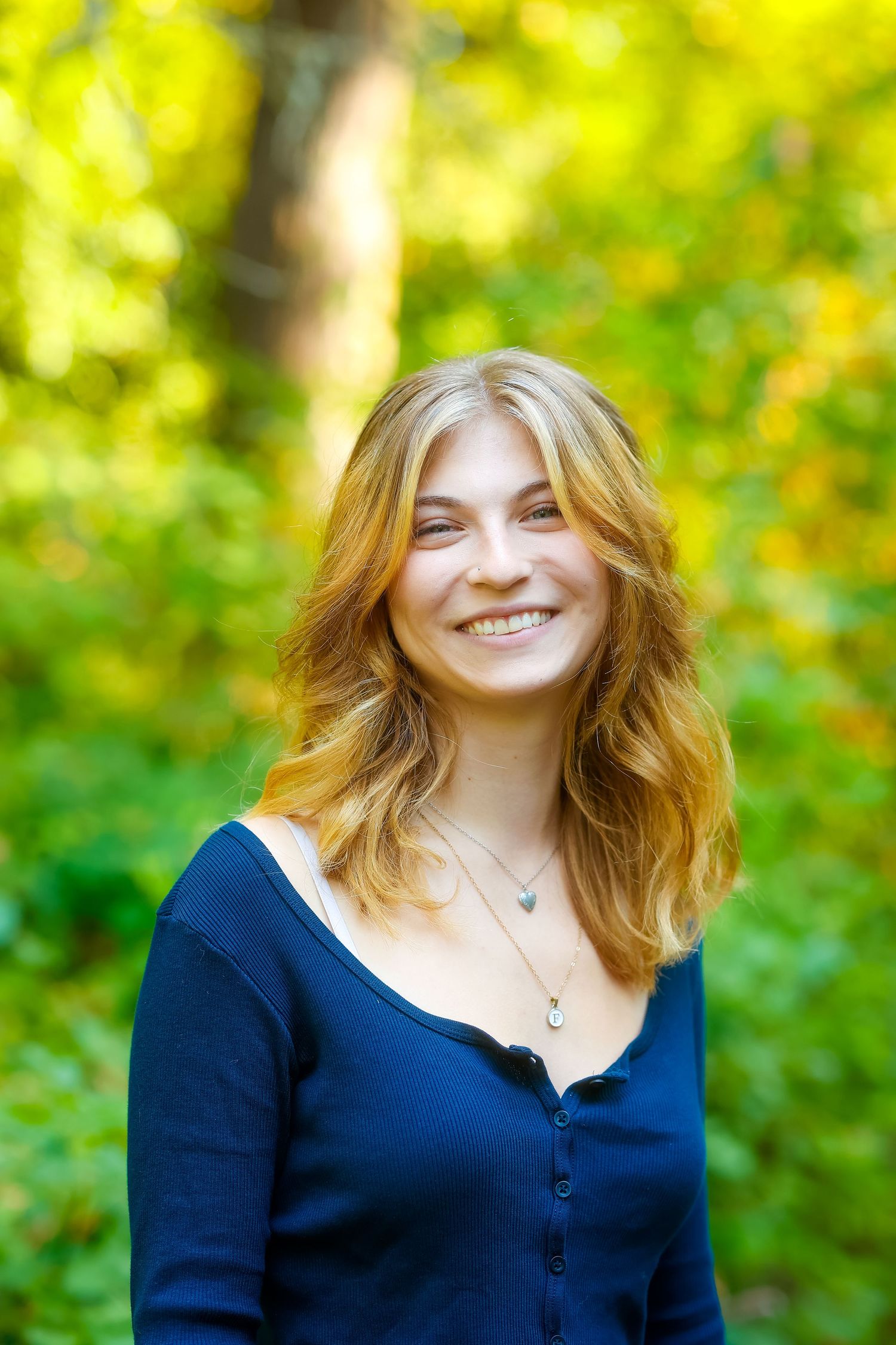 Portrait of someone with wavy blonde hair wearing a navy blue button-up top smiling outdoors with yellow foliage in background.