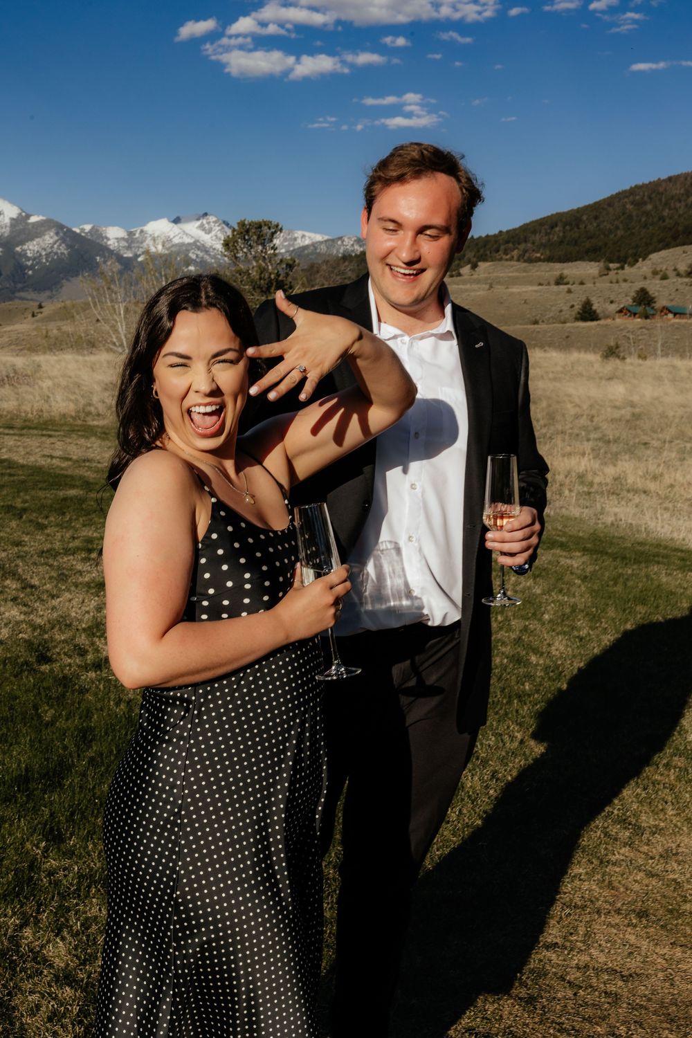 A couple laughs joyfully while holding champagne glasses in an outdoor setting with mountains and blue sky in the background.