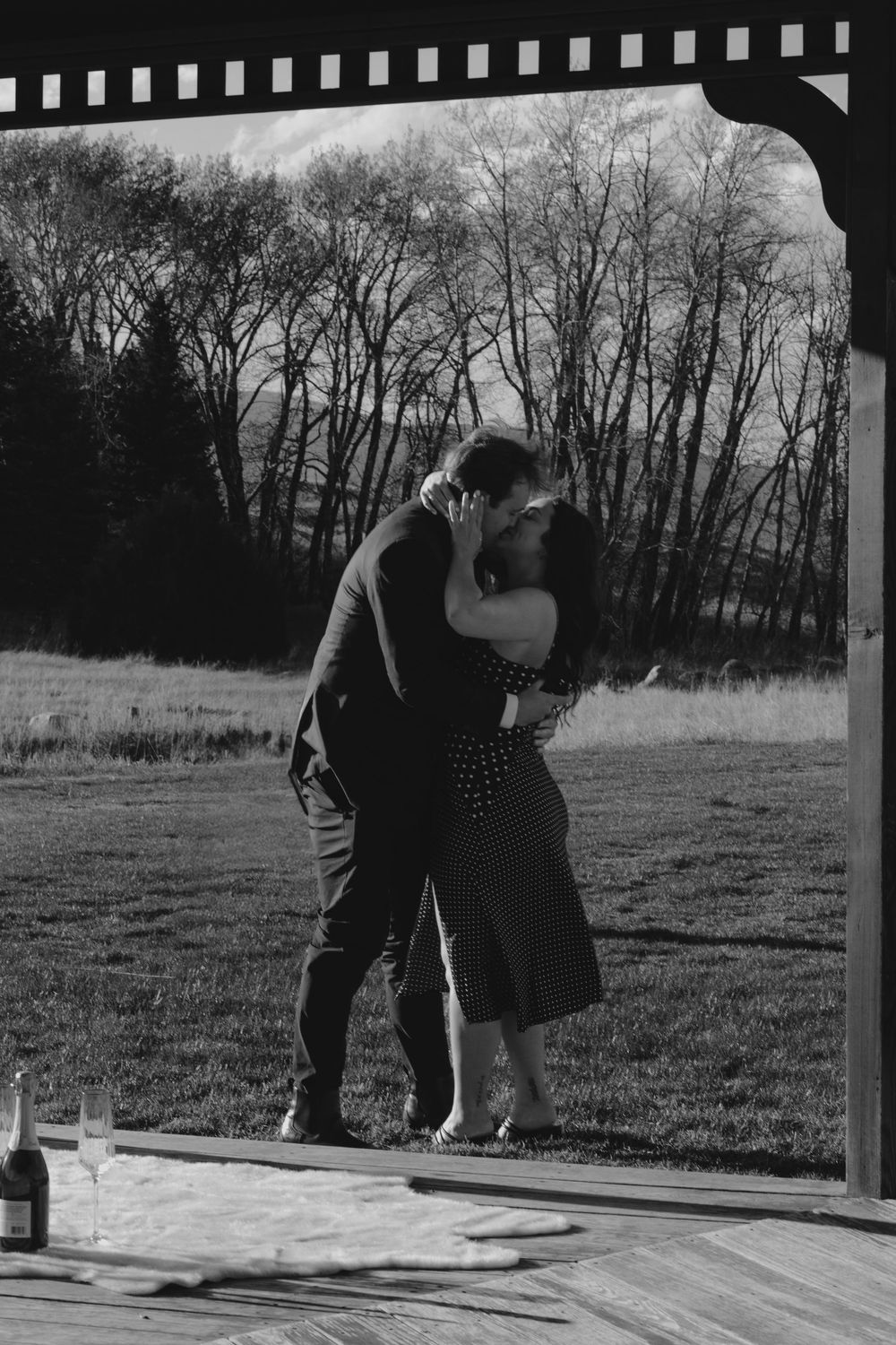 A romantic black and white photo of a couple embracing on a wooden deck with trees in the background.