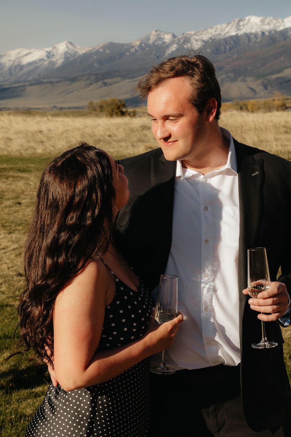 A couple shares a romantic moment in a field with snow-capped mountains in the background at sunset.