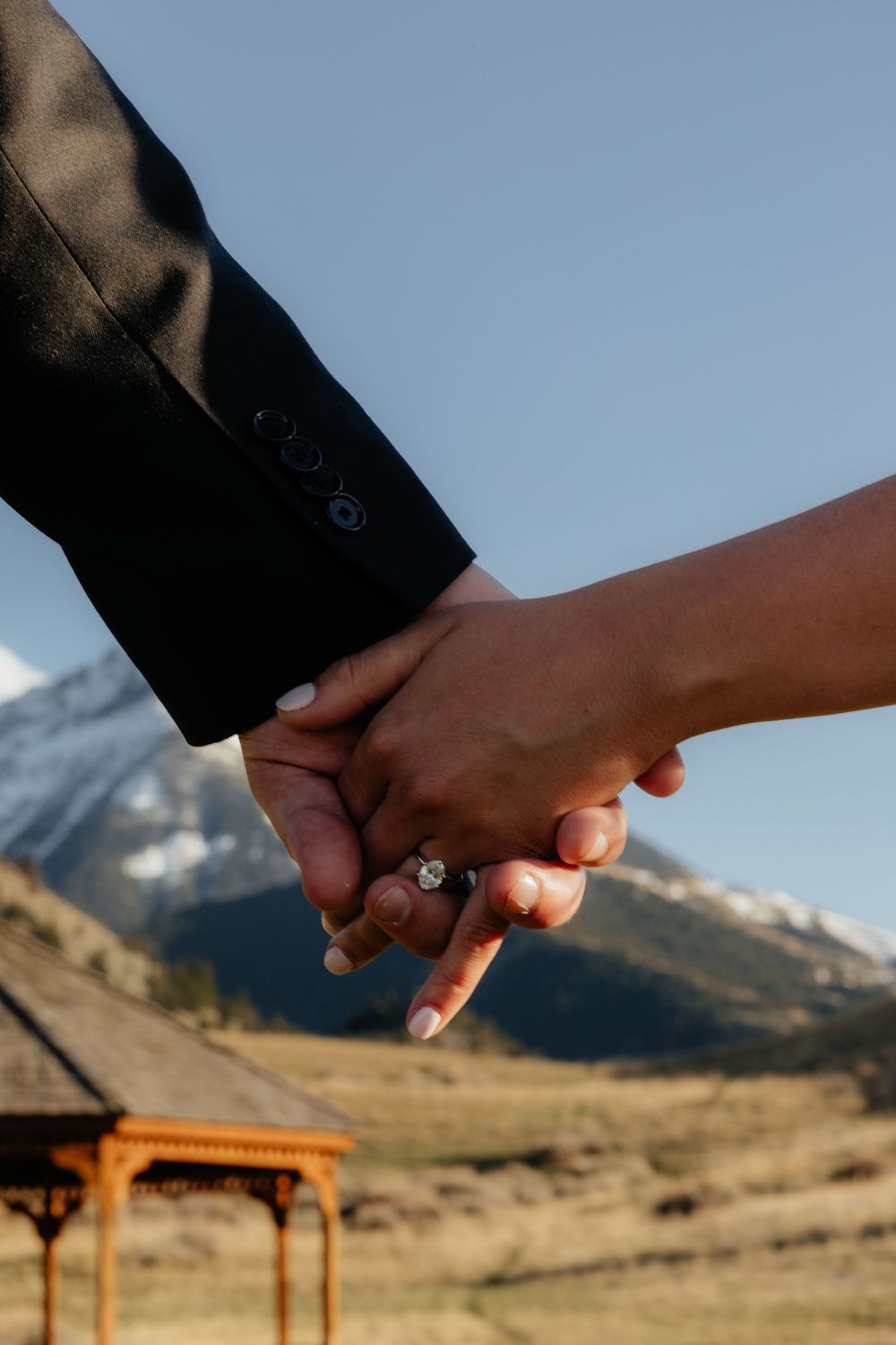 A couple holds hands showing an engagement ring against a scenic mountain landscape with wooden gazebo in background.