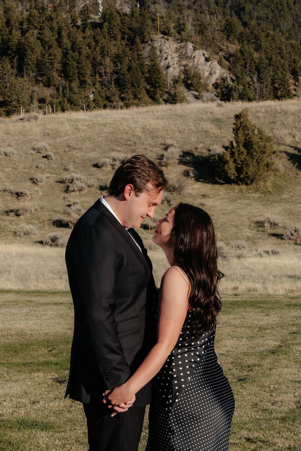 A romantic couple shares intimate moments in a grassy outdoor field with mountains and trees in the background.