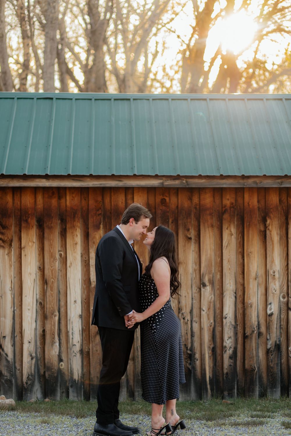 A romantic couple stands close together in front of a rustic wooden barn with a teal metal roof at sunset.