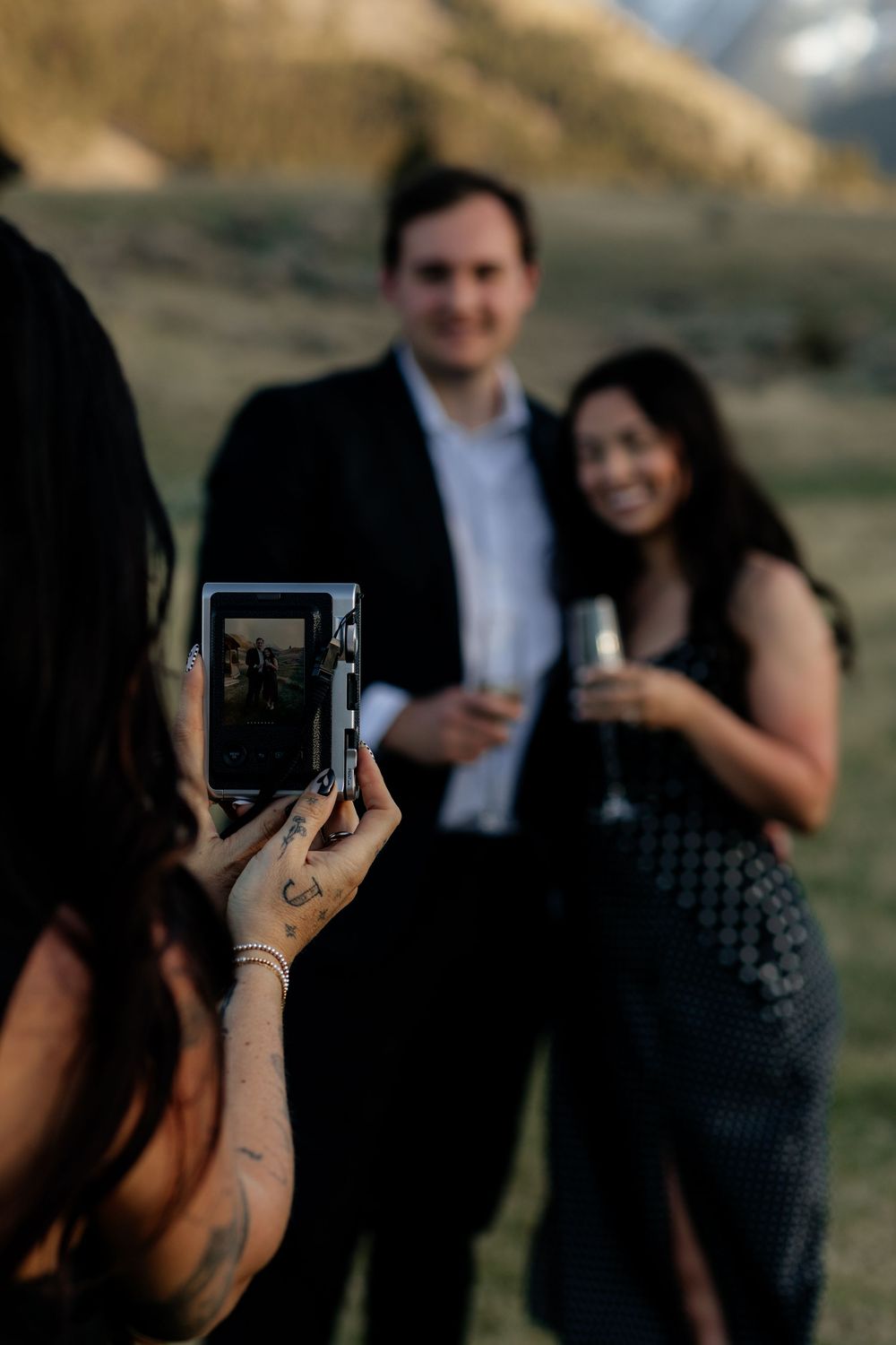 Someone taking a photo on a phone of a couple holding drinks outdoors against a mountain backdrop at dusk.
