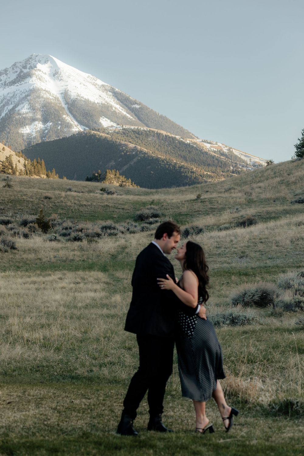 A romantic couple shares intimate moments against a backdrop of snow-capped mountains and rolling hills at golden hour.