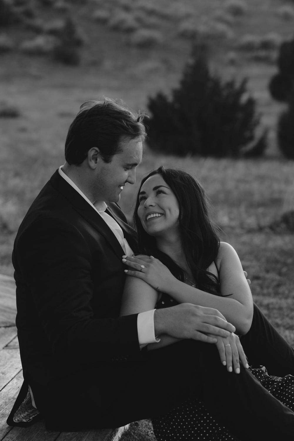 A couple in formal attire share an intimate candid moment while sitting outdoors in black and white photography.